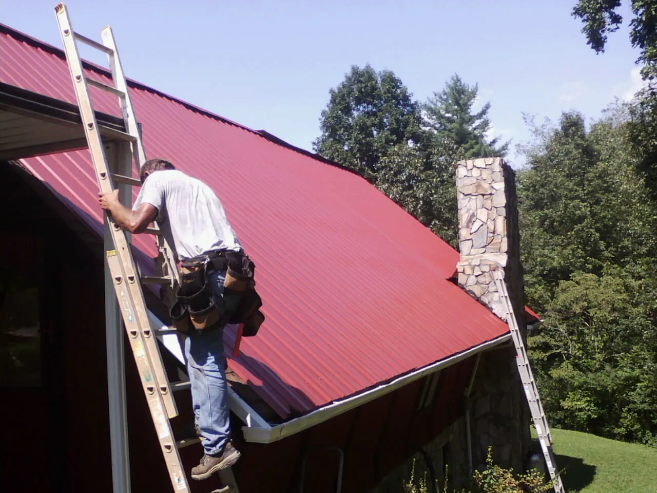 Roofer on a ladder beside a red metal roof and stone chimney — Soffit & Fascia Repair by Roofing Brothers in Alberta